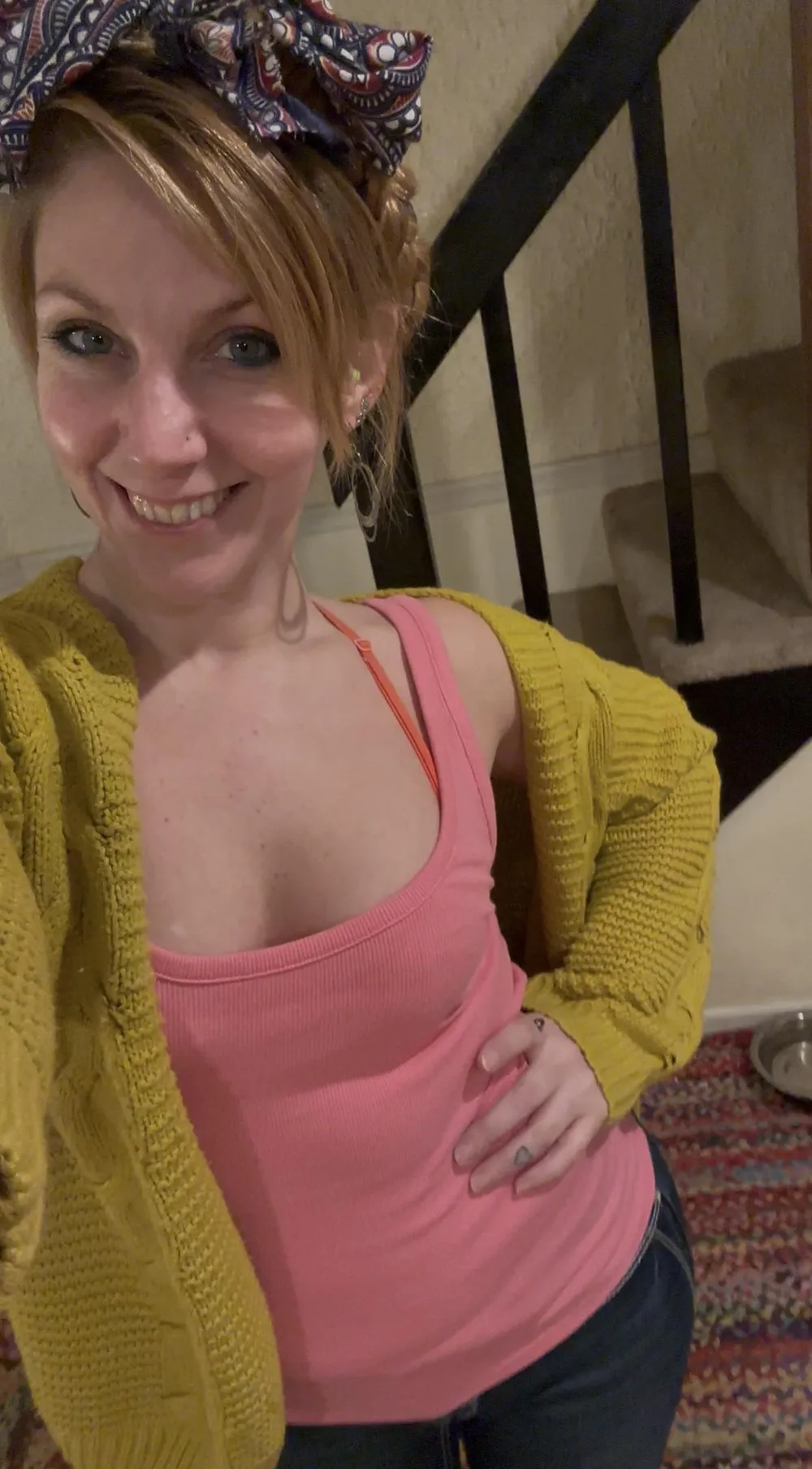 Red-haired OnlyFans model smiling in a pink top and yellow cardigan while taking a selfie indoors near a staircase